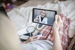 A woman in pajamas is holding a tablet while taking a video call with a doctor from the comfort of her home.