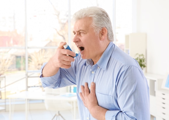 Senior man using an inhaler in a bright room, wearing a blue shirt and expressing discomfort due to asthma.