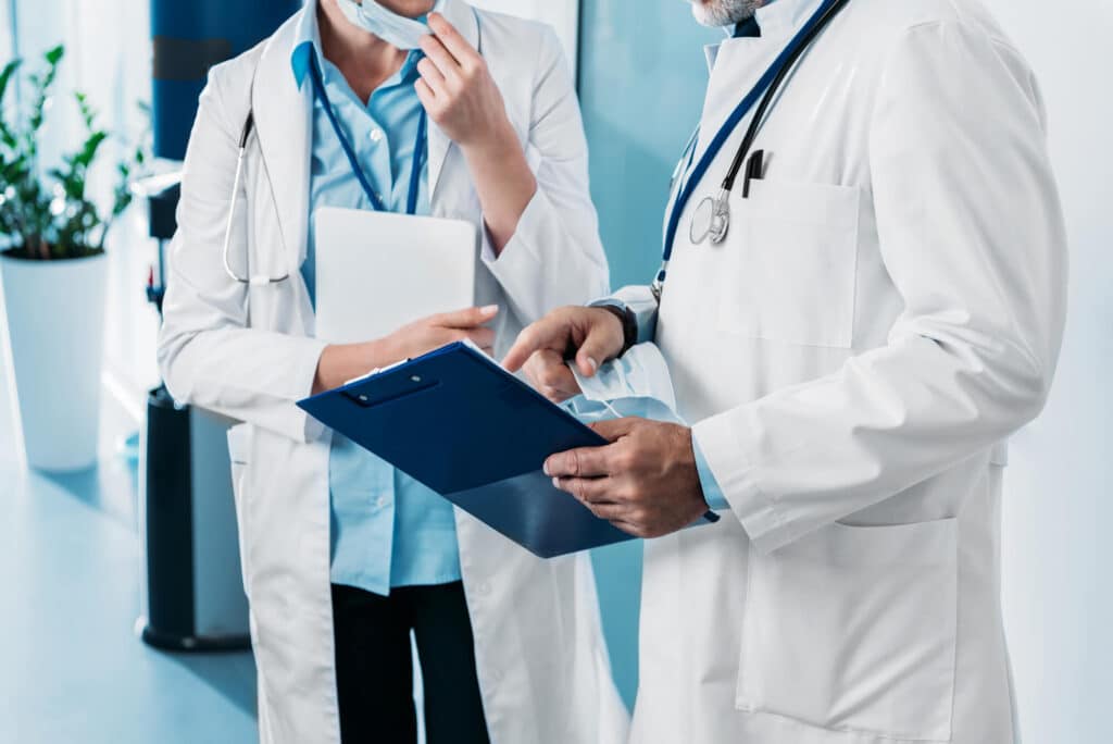 Two medical professionals in white lab coats are discussing over a clipboard in a clinical setting. One is holding a tablet equipped with remote monitoring capabilities, and both have stethoscopes around their necks.