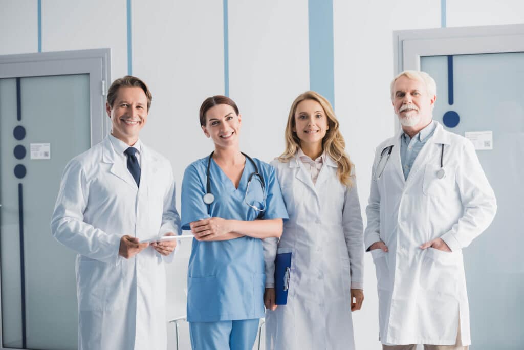 Four medical professionals, two men and two women, stand together in a hospital hallway. They wear white coats and scrubs, smiling while looking at the camera, symbolizing the future of healthcare that includes advancements like remote monitoring.