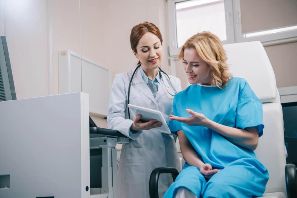A doctor in a white coat shows a tablet displaying remote monitoring data to a seated patient in a blue medical gown, amid medical equipment and a window in the background.