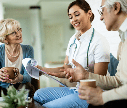 A healthcare professional discusses paperwork with two elderly individuals, who are seated and holding cups while engaging in conversation. This serene and caring scene showcases the compassionate service highlighted on our landing page.