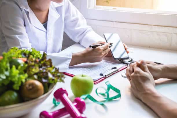 A person in a white coat, possibly one of the RDs, discusses a diet plan with another individual at a table. Nearby are a bowl of vegetables, an apple, pink dumbbells, and a measuring tape.