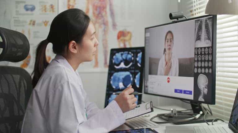 A doctor in a white coat conducts a virtual consultation with a patient via a computer screen, exemplifying the advancements in remote care. X-ray images are visible on another monitor beside her, reflecting upcoming 2025 CMS changes aimed at improving telehealth services.