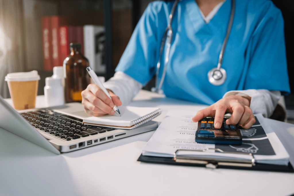 A person in medical scrubs writes notes while using a calculator and laptop at a desk filled with bottles, a notepad, and a coffee cup, seamlessly navigating CPT codes to streamline medical billing.