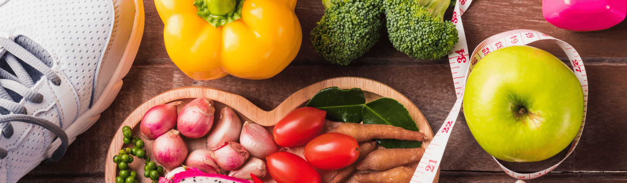 A variety of healthy foods like a yellow bell pepper, broccoli, tomatoes, garlic, an apple, and a Sleep Buddy product with a measuring tape on a wooden surface.