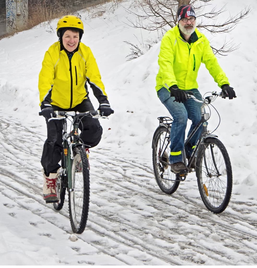 Two people wearing yellow jackets and helmets are enjoying the most wonderful time as they ride bicycles on a snowy path.