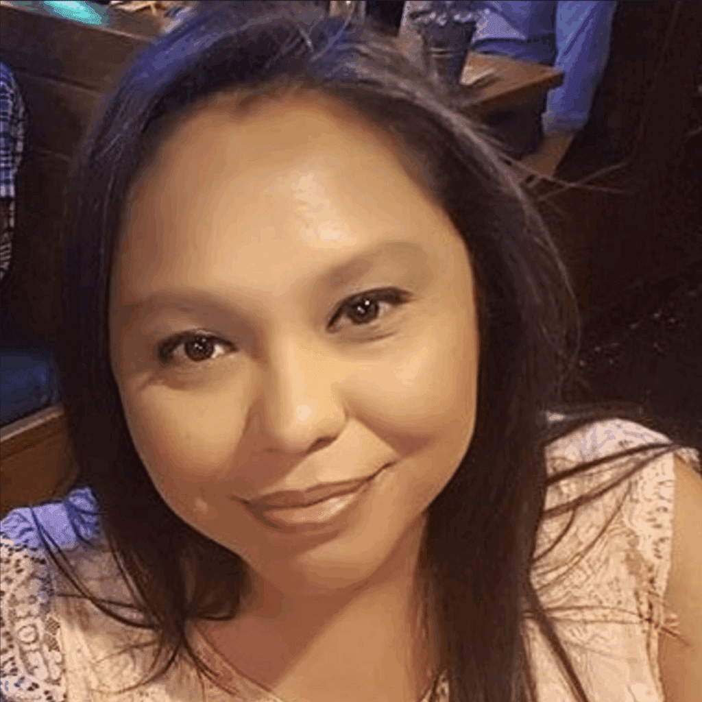 A woman with long dark hair and a light pink lace top smiles at the camera in a restaurant setting, embodying the warmth and confidence often seen in success stories.