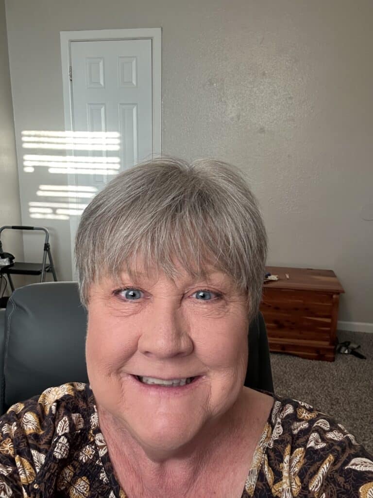 An older woman with short gray hair smiles at the camera in a home office with beige walls and a closed door, embodying the heart of real-life Success Stories.