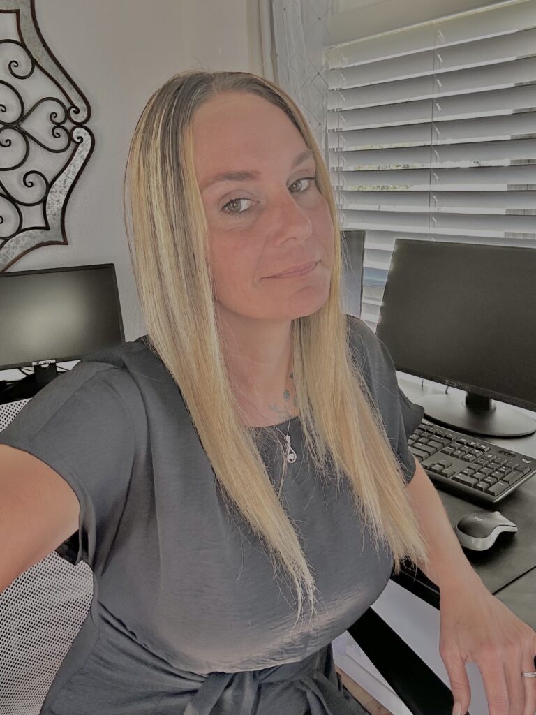 A woman with long blonde hair sits at a desk with two computer monitors and a keyboard, exemplifying the dedication behind many Success Stories, with window blinds in the background.