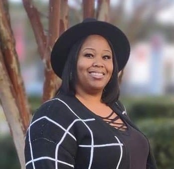A woman wearing a black hat and a black top with a white grid pattern stands outdoors, smiling at the camera—radiating the confidence found in many success stories. Trees and greenery create a vibrant backdrop.
