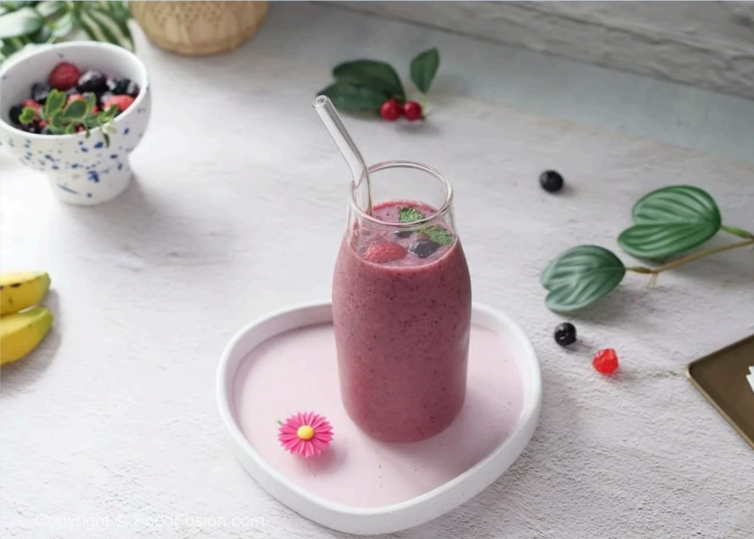 A pink smoothie in a glass with a metal straw, garnished with berries and mint, sits on a heart-shaped tray—a perfect way to kickstart your health-focused New Year's resolution. A small bowl of berries and sliced banana awaits in the background.