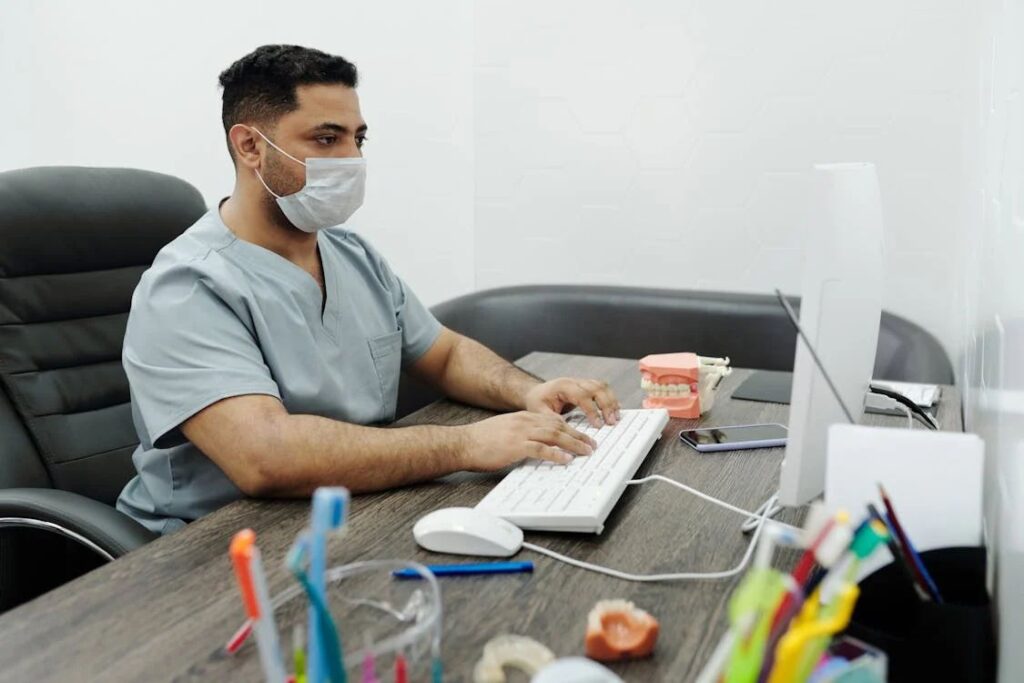 A man in scrubs and a mask sits at a desk, diligently typing on a keyboard. Beside the dental model, phone, and colorful pens lies the Healthcare Provider’s Guide—essential for mastering details like the 99487 CPT Code to maximize reimbursement in the practice.