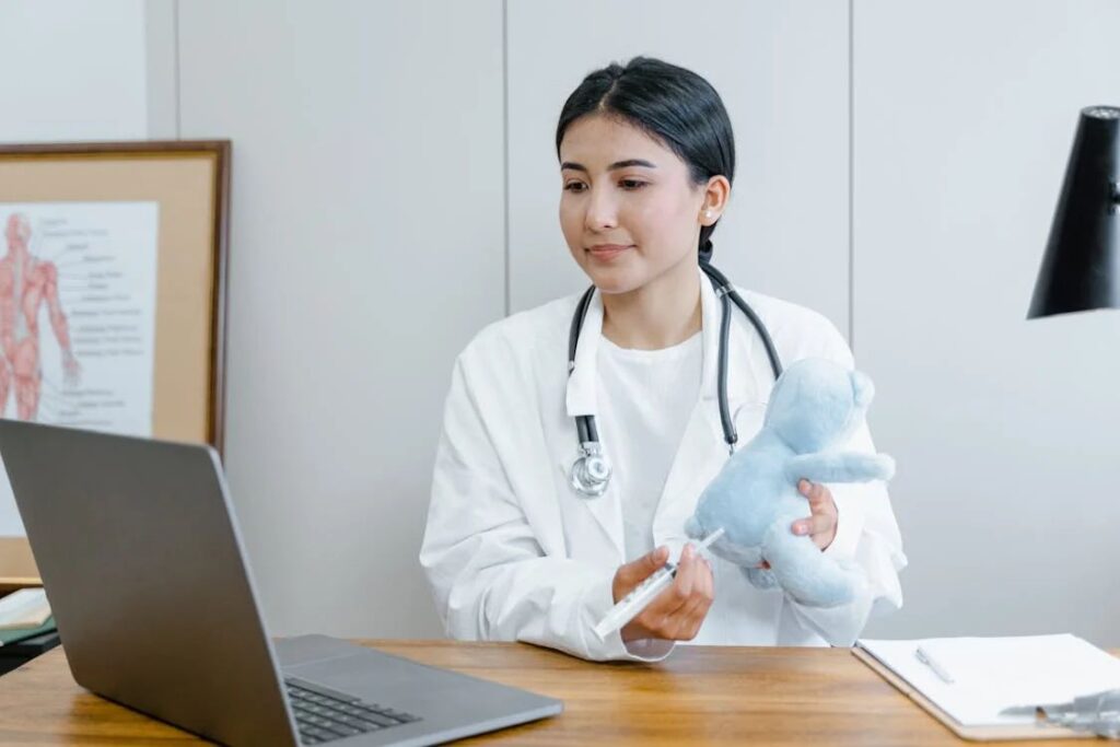 A doctor, focusing on maximizing reimbursement, holds a syringe and a teddy bear while sitting at a desk with a laptop and medical chart.