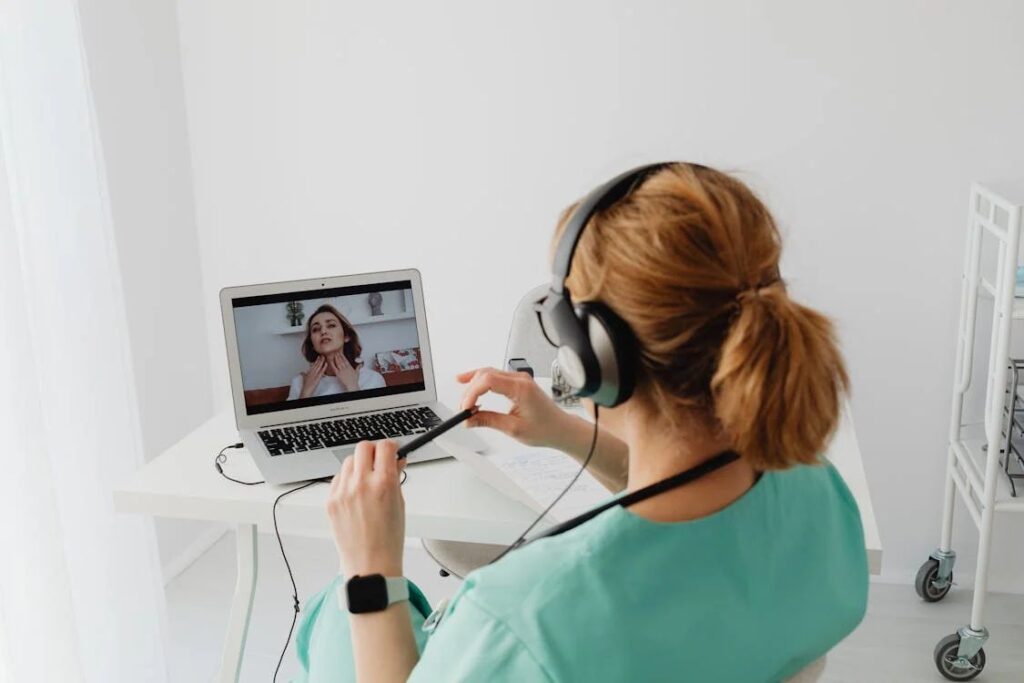 A healthcare professional in green scrubs is engaged in a Remote Patient Monitoring session, video calling a patient on a laptop while wearing headphones, with medical equipment nearby.