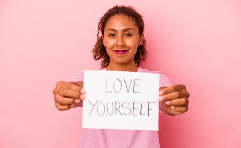 A person stands against a pink background, holding a sign with the handwritten message "LOVE YOURSELF" toward the camera, inspiring self-love—perfect for a patient newsletter feature in March 2025.