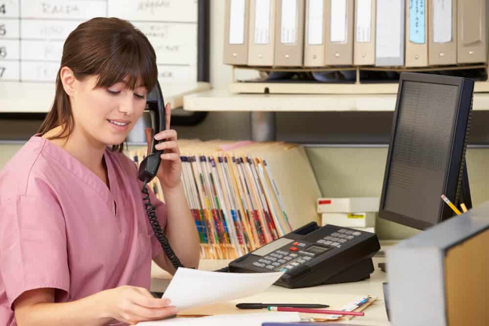 A woman in pink scrubs sits at a desk, talking on the phone while reviewing documents related to Non-Complex CCM, with folders and office supplies organized around her.
