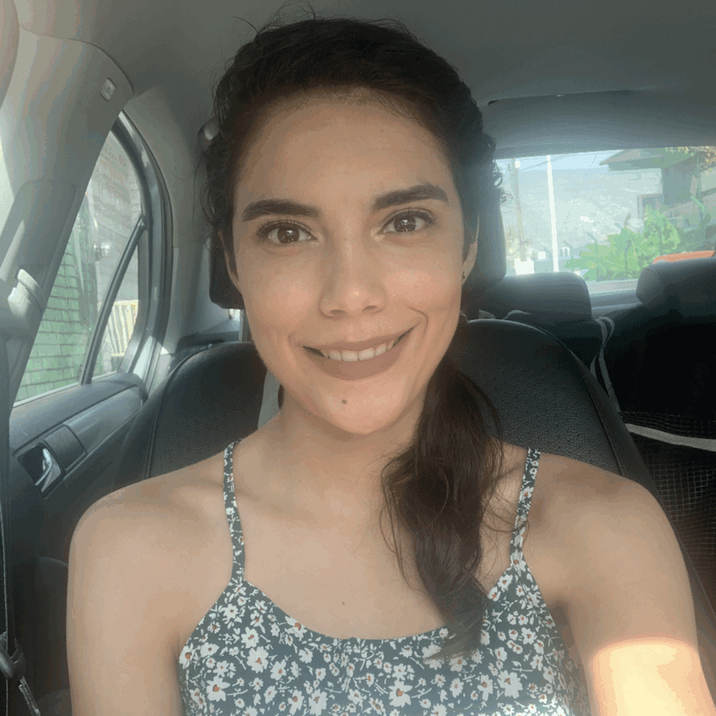A woman with dark hair in a side ponytail, wearing a floral sleeveless top, sits in the driver’s seat of a car and smiles at the camera—sunlight streams in, capturing a moment from her inspiring weight loss success story.