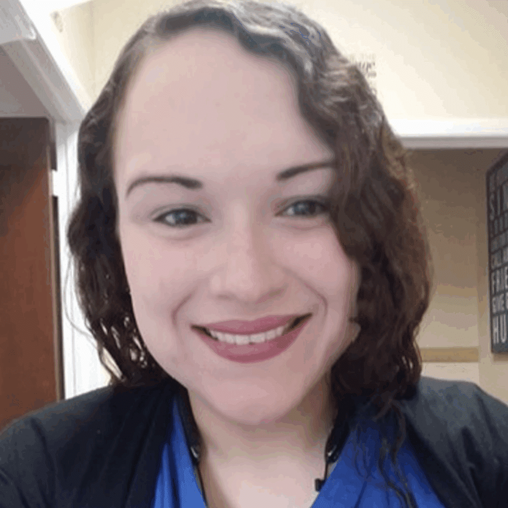 A person with wavy brown hair and light skin smiles at the camera, wearing a blue top and black sweater, standing indoors near a doorway—radiating confidence often seen in weight loss success stories.