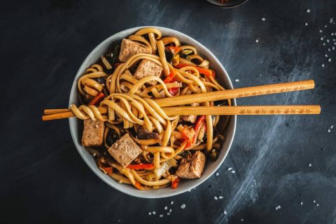 A bowl of stir-fried noodles with tofu and vegetables, ideal for weight loss, with chopsticks resting on top, on a dark textured surface.