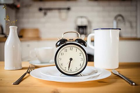 A vintage black alarm clock centered on a white plate, with a cup, spoon, and milk bottle in a diet-conscious kitchen setting.