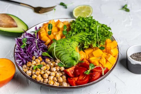 A colorful, weight-loss-friendly salad with sliced avocado, mango, cherry tomatoes, purple cabbage, roasted chickpeas, and lettuce, garnished with sprouts. An avocado half and lime are in the background.